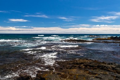 rocky shoreline at black sand beach (punaluu), on the big island of hawaii. blue pacific ocean washing against volcanic rocky shore; blue sky with clouds in the distance. 
