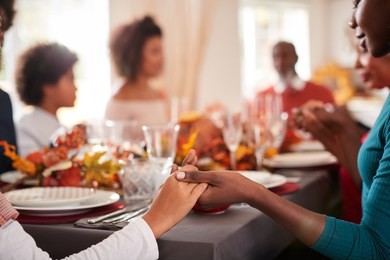 young black adult woman and her daughter holding hands and saying grace with their multi generation family at the thanksgiving dinner table, detail, focus on foreground
