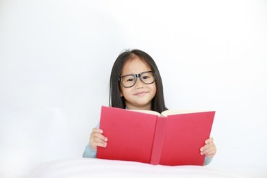 beautiful happy little asian kid girl reading hardcover book lying on bed against white background.