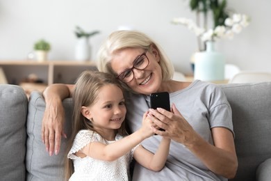 happy grandmother and cute granddaughter using cellphone together, smiling older grandma and child girl having fun taking selfie on phone, cheerful granny with little kid play making photo on mobile