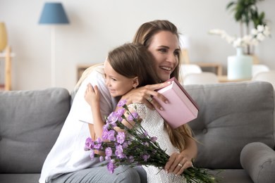 grateful loving young mom thanking embracing little cute daughter holding gift box and flowers bouquet, kid girl hugging happy mommy congratulating mum with birthday or mothers day making present