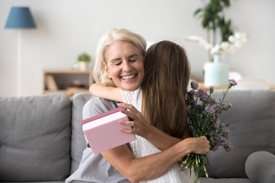 happy senior grandma hugging granddaughter thanking for present holding flower bouquet, smiling excited old grandmother embracing little grandchild girl congratulating granny giving birthday gift box