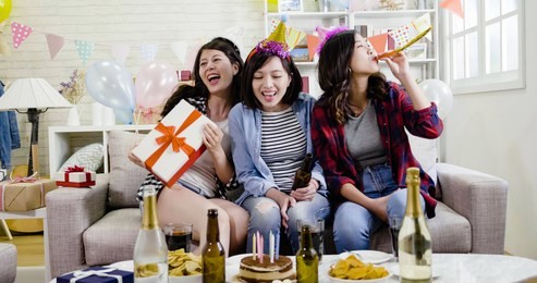 group of friends enjoy birthday party in daytime. young girls playing whistle and sending present for surprised celebrating 20s. asian women having fun at home celebration lifestyle concept.