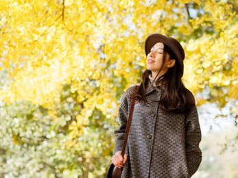 portrait of young chinese woman in fashionable coat and hat with golden autumn forest background in park, carring a bag. cute girl in good mood posing in autumn day, enjoying good weather.