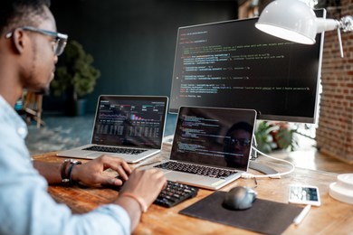 young african male programmer writing program code sitting at the workplace with three monitors in the office. image focused on the screen