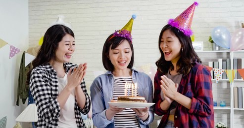 three cute ladies holding lighted cake with candles on fire cheerfully singing songs and clapping hands. young girls with party hats celebrating birthday at home. decorated house for surprise.