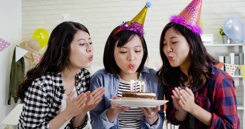 three cute ladies holding lighted cake blowing fire on candles together celebrating birthday house party. young asians with funny hats finished making wishes. confident friends clapping hands.