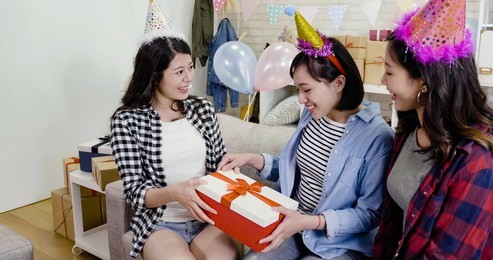 cheerful sisters giving present to girl for congratulating her 20s birthday in the decorated house. young best friends celebrating home party wearing funny hats sitting on couch in living room.