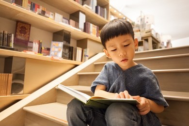 portrait shot of an asian little boy is sitting on the stairs, reading book in bookstore paying full attention