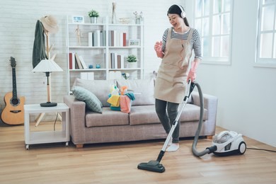 smiling excited young housewife havig fun while cleaning floor with vacuum cleaner. happy woman doing housework at home enjoy music wearing earphones. asian lady in arpon singing dancing house chores