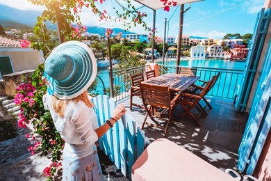female tourist with blue sun hat staying in assos village in front of cozy veranda and admiring turquoise colored bay of mediterranean sea and beautiful colorful houses of in kefalonia, greece