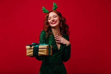 lovable european girl with christmas present expressing happiness. refined ginger young woman in green dress laughing on red background in new year.