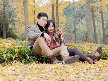 asian couple in love sits on fallen leaves in park. young family and autumn concept. man and woman with smiling faces on autumn trees background. girl and boy or happy lovers on a date.