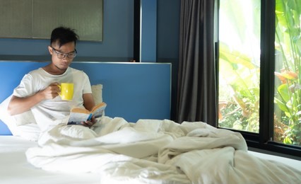asian business man have beard sit reading the book on the bed in the morning, at the same time drink coffee. concept working on the bed in the morning.