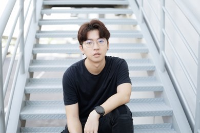 handsome asian casual glasses man sit on a staircase and smile posing on gray background.
portrait of young korean against staircase background.