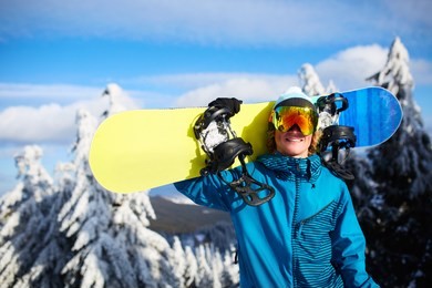 smiling snowboarder posing carrying snowboard on shoulder at ski resort near forest before backcountry freeride and wearing reflective goggles, colorful fashion outfit. modern snowboarding equipment.
