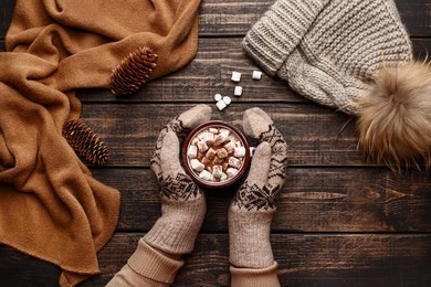 winter flat lay with scarf, knitted hat and female hands in mitten holding mug with hot chocolate or cocoa with marshmallow on dark rustic wooden table. christmas and new year background. top view