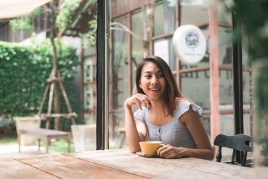 cheerful asian young woman drinking warm coffee or tea enjoying it while sitting in cafe. attractive happy asian woman holding a cup of coffee.