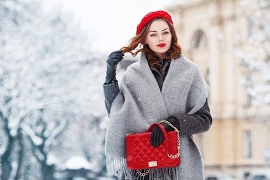 outdoor portrait of young beautiful  fashionable woman wearing stylish winter gray coat, woolen scarf, red beret, leather gloves, holding quilted bag, posing in street of european city. copy space
 