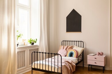 cabinet next to bed in bright bedroom interior with black poster and plants on window sill. real photo