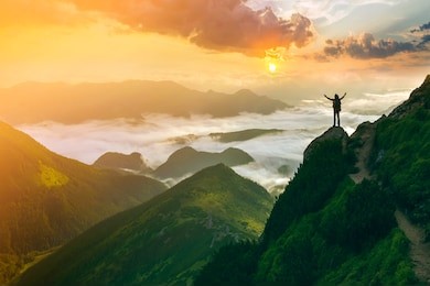 wide mountain panorama. small silhouette of tourist with backpack on rocky mountain slope with raised hands over valley covered with white puffy clouds. beauty of nature, tourism and traveling concept