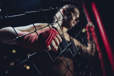 mma is young girl in octagon during fights without rules gaining strength at grid of boxing ring, hard clutching links fist wrapped red protective bandage. selective focus