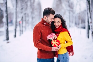celebration of st valentine's day. happy and beautiful young couple in love are walking together outdoors in winter city park and hugging and kissing each other. gifts and flowers