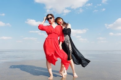 happy smiling pretty chinese girls having fun on sunny beach. stylishly dressed in red dress and black dress, sunglasses, best young friends hand in hand and hug in love. 