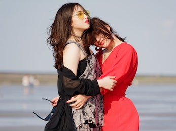happy smiling pretty chinese girls having fun on sunny beach. stylishly dressed in red dress and black dress, sunglasses, best young friends hand in hand and hug in love. 