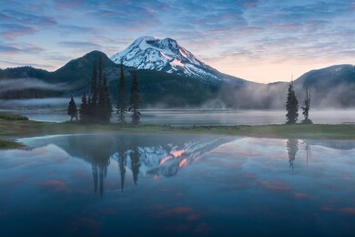 south sister and broken top reflect over the calm waters of sparks lake at sunrise  in the cascades range in central oregon, usa in an early morning light. morning mist rises from lake into trees.