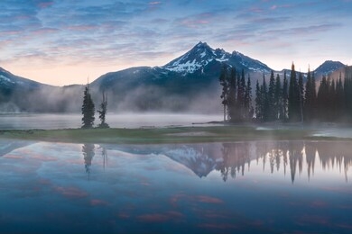 south sister and broken top reflect over the calm waters of sparks lake at sunrise  in the cascades range in central oregon, usa in an early morning light. morning mist rises from lake into trees.