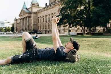 a young guy with a backpack or a student is lying on the grass during a break or is resting and using a tablet. he is watching a video or reading or something else.