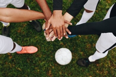 top view of hands of players placed one over the other standing in a huddle. players standing in a huddle joining their hands together in the centre with a soccer ball on the ground.