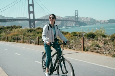 young tourist woman sunglasses relaxing and enjoying the sun riding bike tour in usa. sport girl cycling sightseeing visiting golden state bridge on coast. asian lady ride bicycle on road sunny day