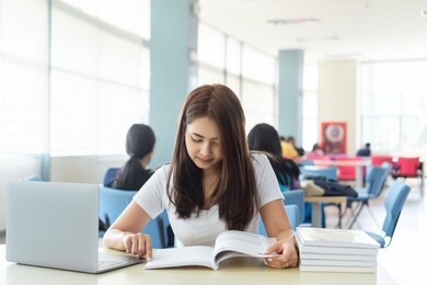 portrait of young asian female student college is reading book while sitting on table with laptop and stack of textbooks with blurred background of people in library. education and learning concepts.