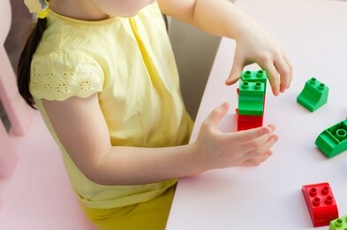 little girl in a yellow shirt playing with lego/construction toy blocks/colorful plastic bricks at sunny room at home. top view