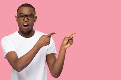 surprised black man pointing fingers at copy space looking at camera, amazed african american guy feeling astonished excited advertising product or service isolated on pink blank studio background