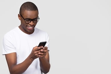 smiling black young man in glasses holding phone looking at smartphone isolated on white grey studio background with copy space aside, happy african guy using mobile applications texting on cellphone