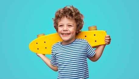 charming curly boy holding yellow pennyboard and looking away isolated on aqua blue background.
