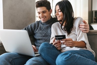 happy young couple sitting on a couch at home, using laptop computer, showing credit card