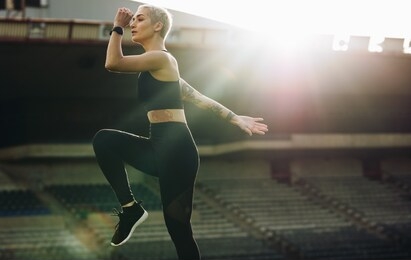 woman doing high knee lifts in a stadium with sun in the background. woman in fitness clothes doing work out in a track and field stadium.