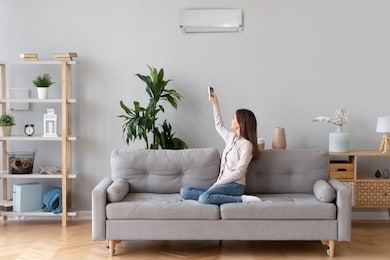 young happy woman switching on air conditioner sitting on couch at convenient cozy home, lady relaxing on sofa in living room holding remote climate control to cooler system set comfort temperature