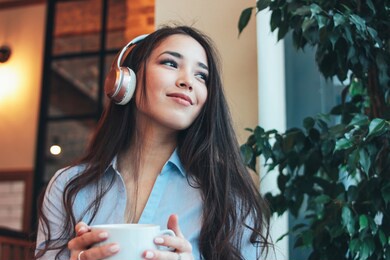 beautiful charming brunette smiling asian girl enjoys music in headphones with cup of coffee or tea in cafe