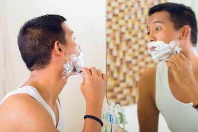 asian man shaving in front of bathroom mirror