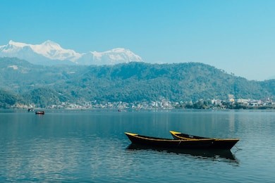 two empty boats floating on the surface of phewa lake, pokhara, nepal. in the back more boats with tourists. cityscape on the shore. high himalayan peaks in the back. chilled and relaxed atmosphere.
