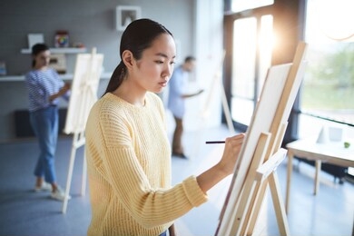 young asian woman in yellow sweater making sketch on paper while standing in front of easel at lesson of painting