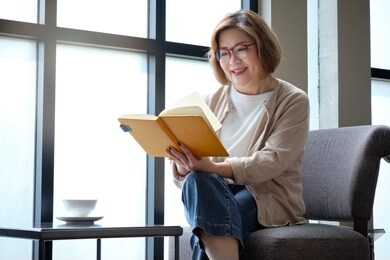 elderly woman reading a book by the window on relaxing day with a cup of coffee.