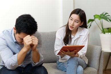 female professional psychologist conducting a consultation to stressful male patient sitting on sofa in her office. she is writing down on a paper and looking at him. psychological consultant concept.