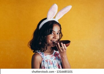 cute little child wearing bunny ears on easter day on color background. girl eating chocolate easter egg