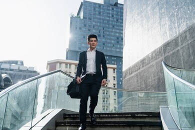 full length of smart asian entrepreneur walking downstairs outdoor, businessman carrying a black bag on shoulder.handsome business hurrying in the urban city .